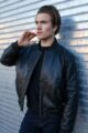 A man wearing a black leather bomber jacket, standing in front of a metallic background and adjusting his collar.