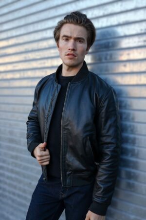 A man wearing the OVLAN Black Leather Motorcycle Jacket, posing confidently against a metallic backdrop. The jacket features a sleek and minimalist design.