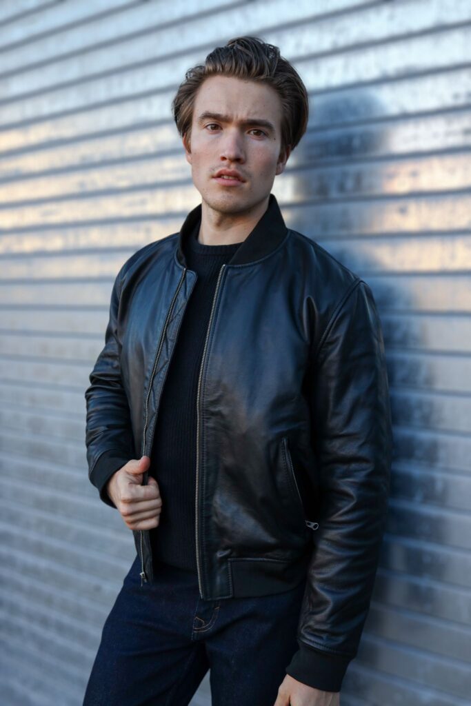 A man wearing the OVLAN Black Leather Motorcycle Jacket, posing confidently against a metallic backdrop. The jacket features a sleek and minimalist design.