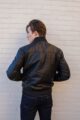 A man modeling the OVLAN black leather motorcycle jacket, leaning against a white brick wall with hands in pockets, showcasing the jacket's sleek and modern style.