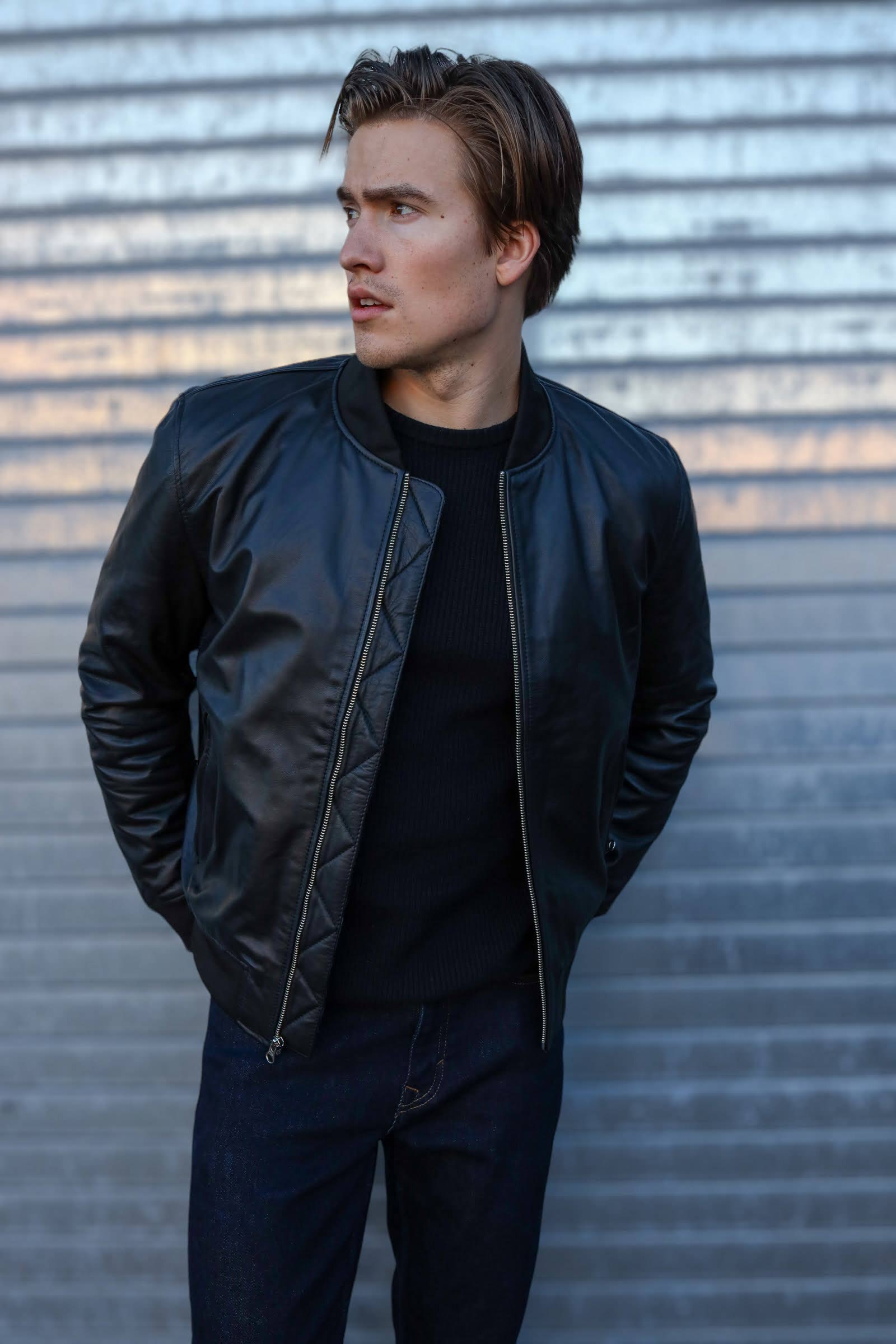 A man wearing a black leather bomber jacket with a quilted pattern on the shoulders, standing in front of a metal background.