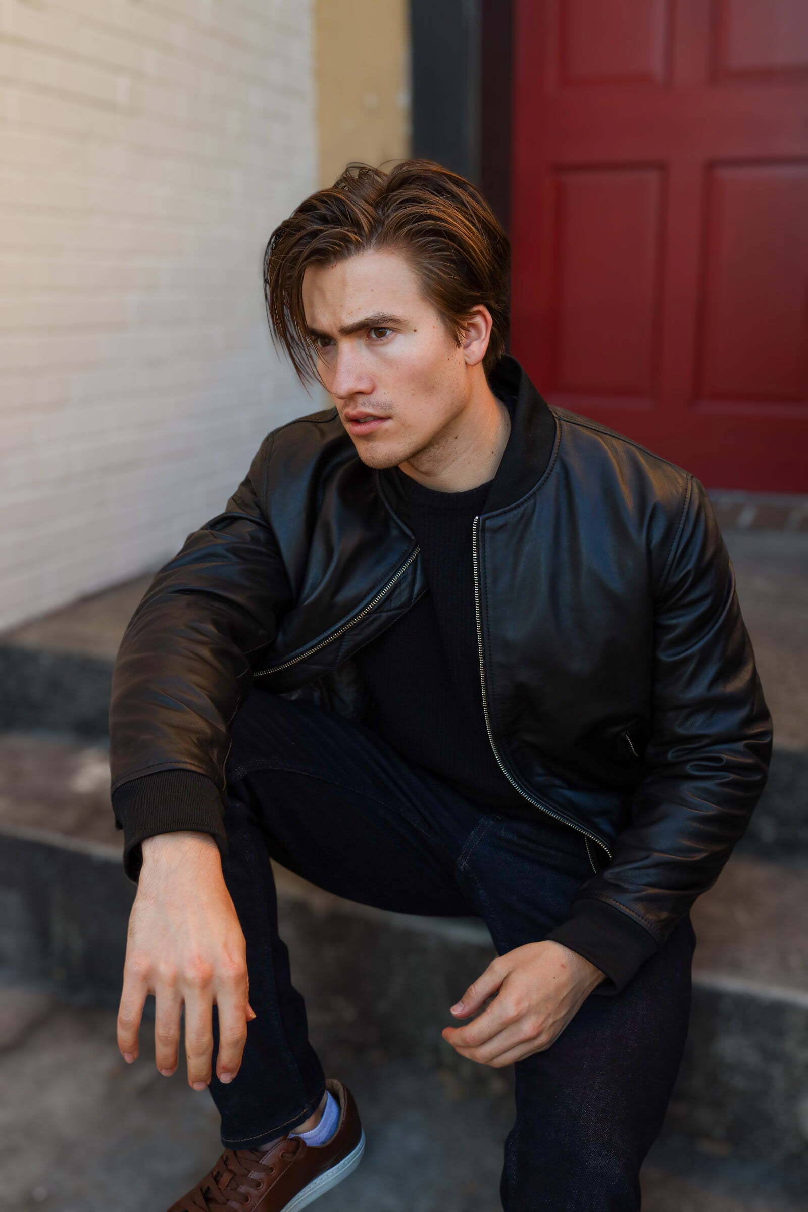 A man crouching on steps, wearing a black leather bomber jacket and dark jeans, with a serious expression.