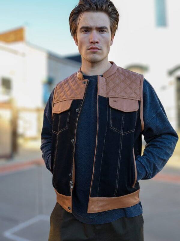 A man wearing an OVLAN denim and leather motorcycle vest with quilted brown leather accents, paired with a gray long-sleeve shirt, standing against a white brick wall.