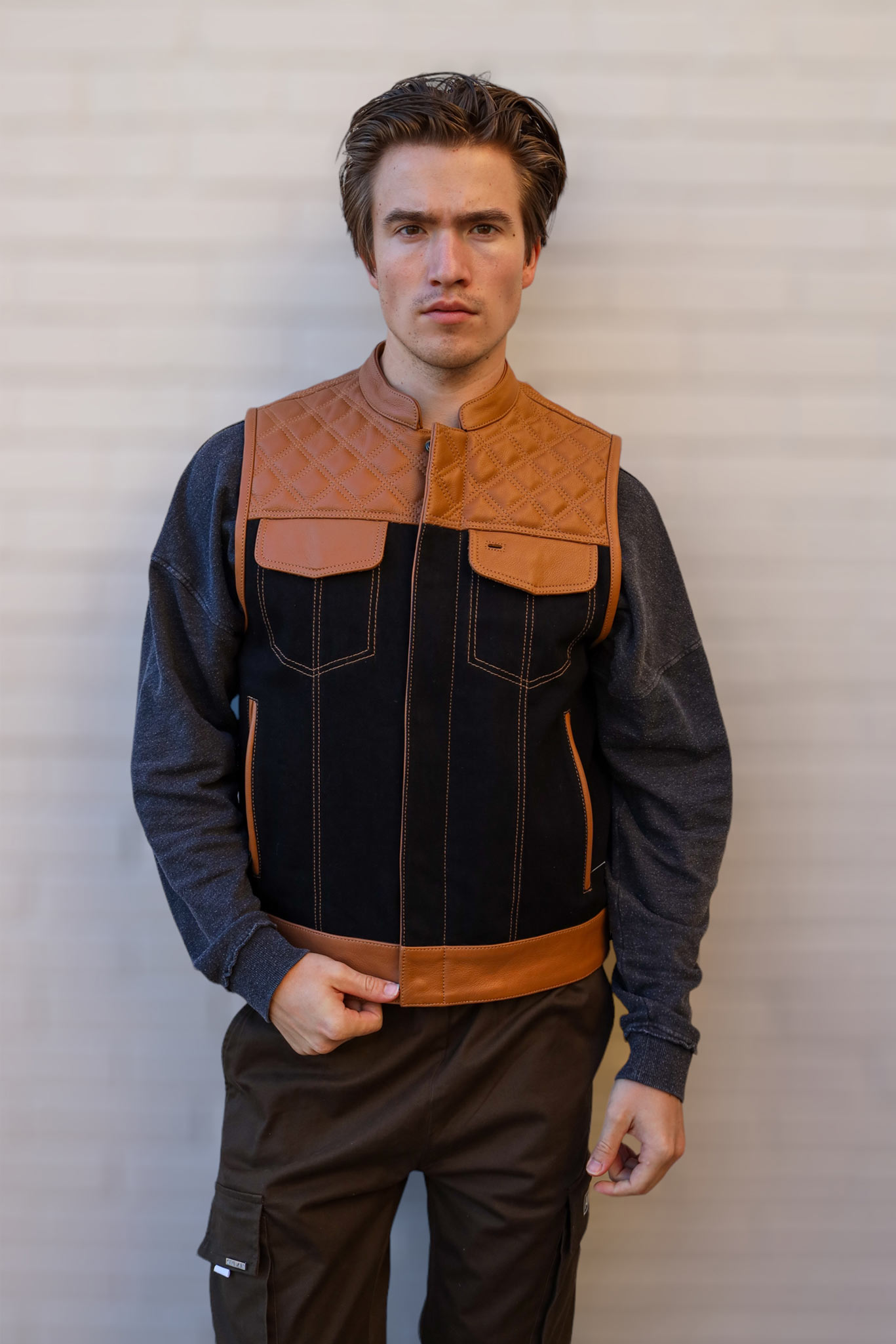 A man wearing an OVLAN denim and leather motorcycle vest with quilted brown leather accents, paired with a gray long-sleeve shirt, standing against a white brick wall.