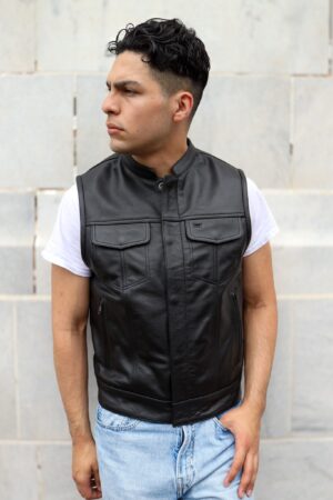 A young man wearing a black leather vest over a white t-shirt, standing in front of a stone wall.