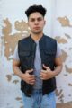 A young man wearing a black leather vest over a gray shirt, standing against a wall with peeling paint.