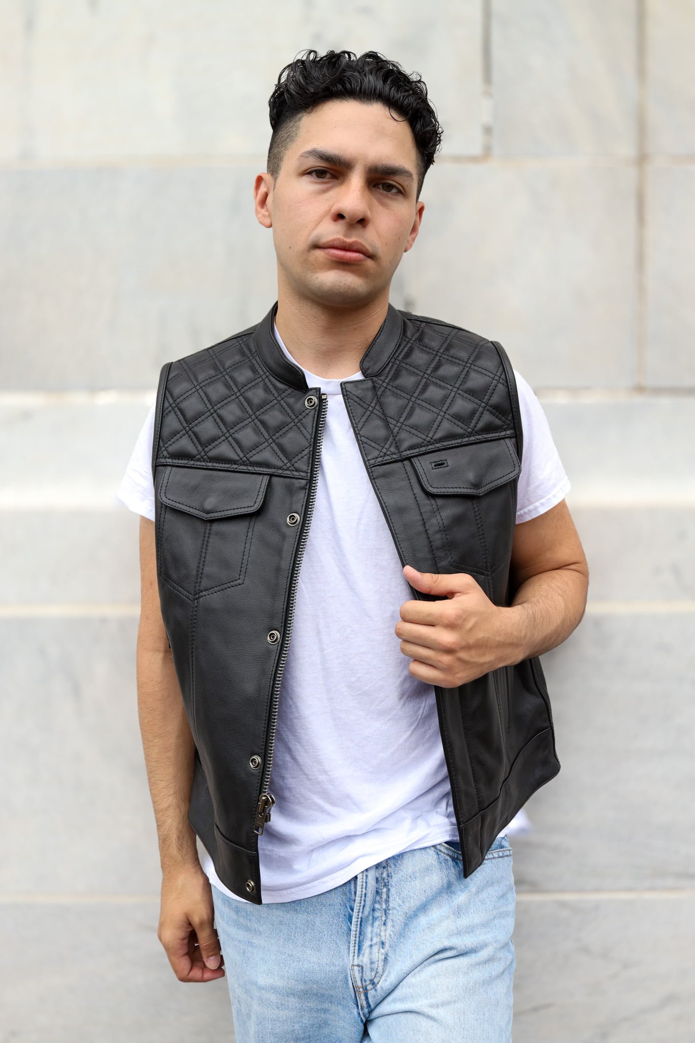 Man modeling a black leather motorcycle vest with diamond stitching and snap-button front pockets, paired with a white shirt and light blue jeans against a neutral background.