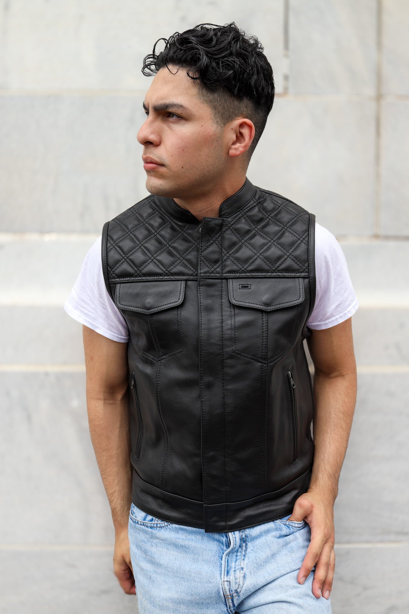 Man wearing an OVLAN black leather motorcycle vest with diamond stitching, paired with a white shirt and light blue jeans, showcasing a stylish side profile.