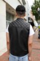The back view of a black leather motorcycle vest with quilted diamond stitching on the shoulders, modeled outdoors on a city sidewalk for a stylish display.