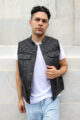 Man modeling a black leather motorcycle vest with diamond stitching and snap-button front pockets, paired with a white shirt and light blue jeans against a neutral background.