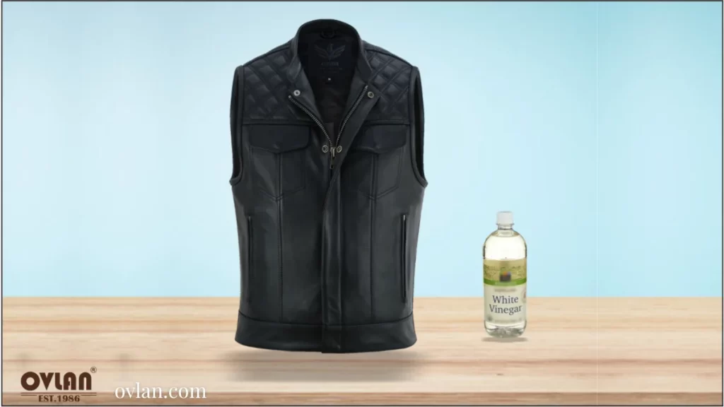 A black leather vest displayed on a table next to a bottle of white vinegar, with a light blue background.