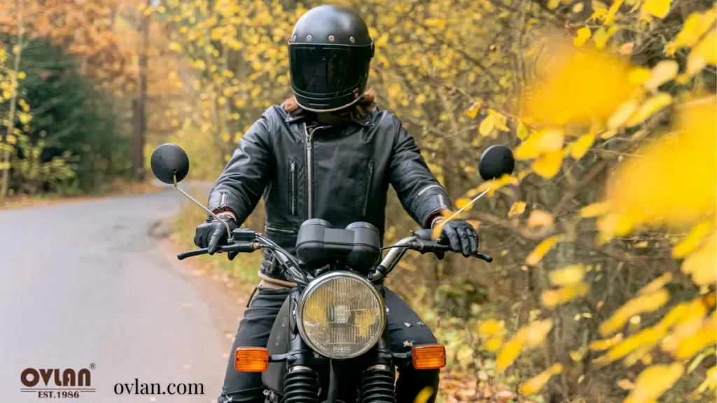 A person riding a motorcycle on a forest road during autumn with yellow leaves in the background, wearing a black helmet, leather jacket, and gloves.
