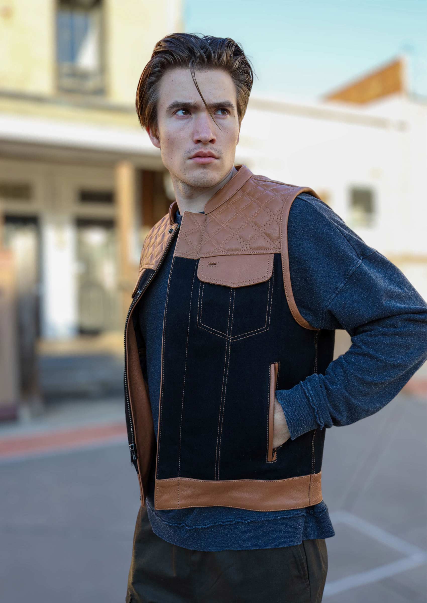 A man confidently wearing an OVLAN denim and leather motorcycle vest with quilted brown leather accents, photographed outdoors with a clear blue sky and urban backdrop.