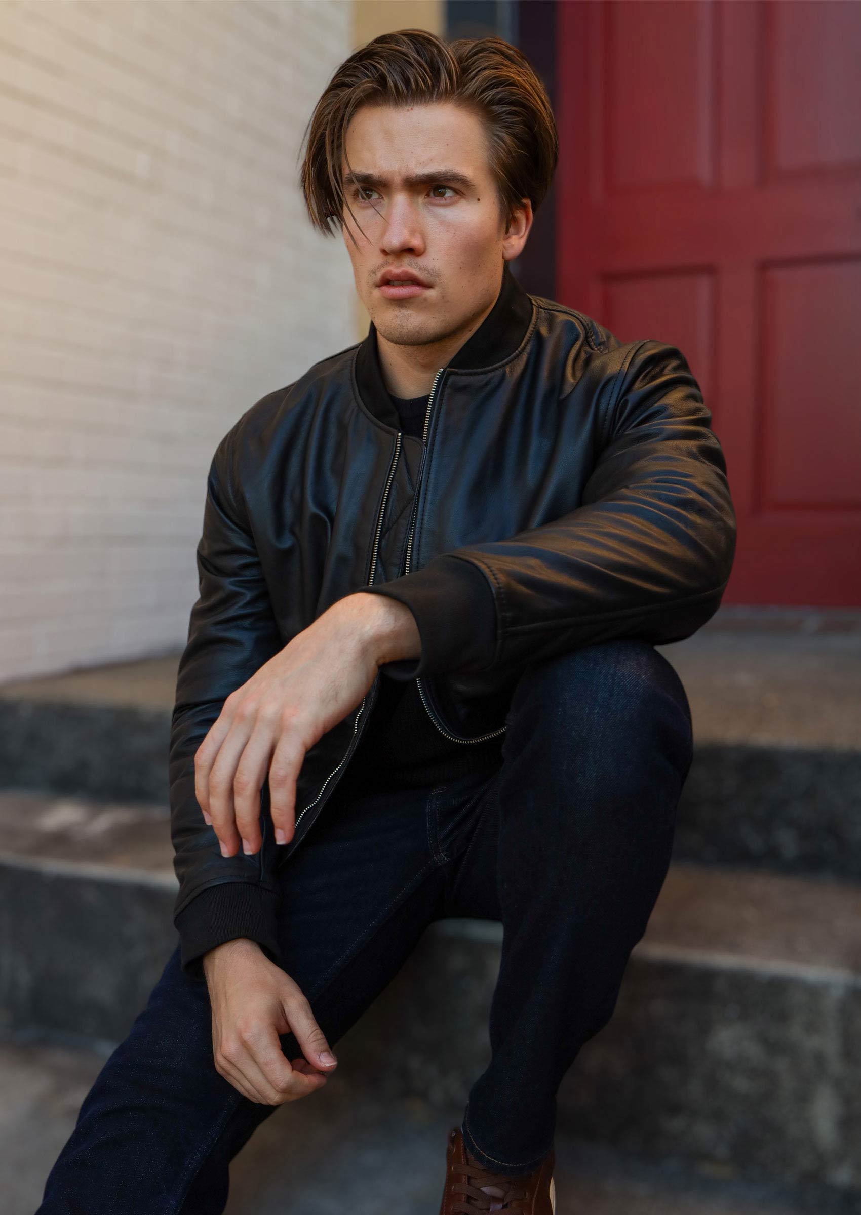 A man in a black leather bomber jacket crouches on stone steps, looking intense with a serious expression. A red door is visible in the background.