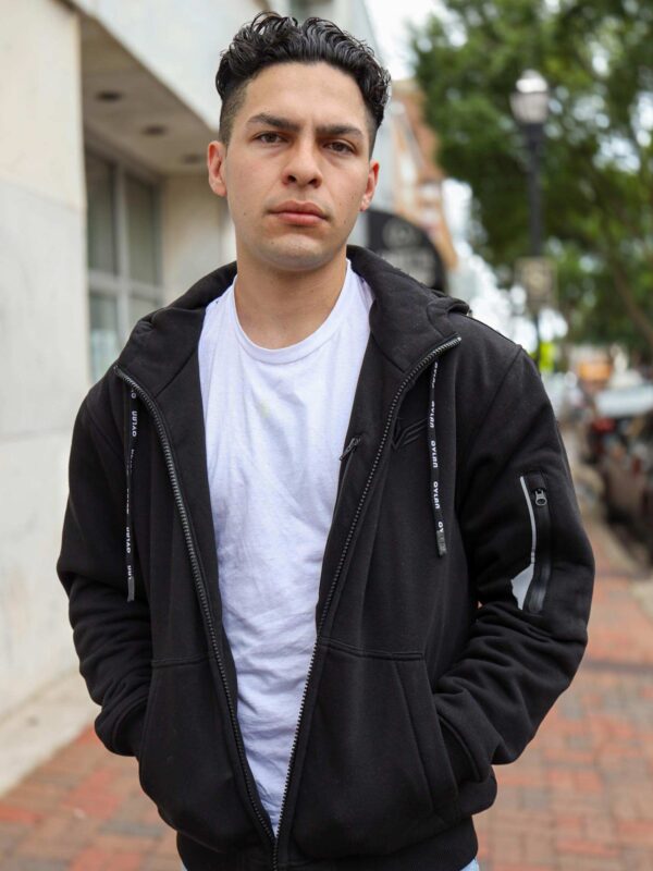 A man wearing a black hoodie with a white t-shirt underneath stands outside, with his hands in his pockets. He has short, curly hair and a serious expression.