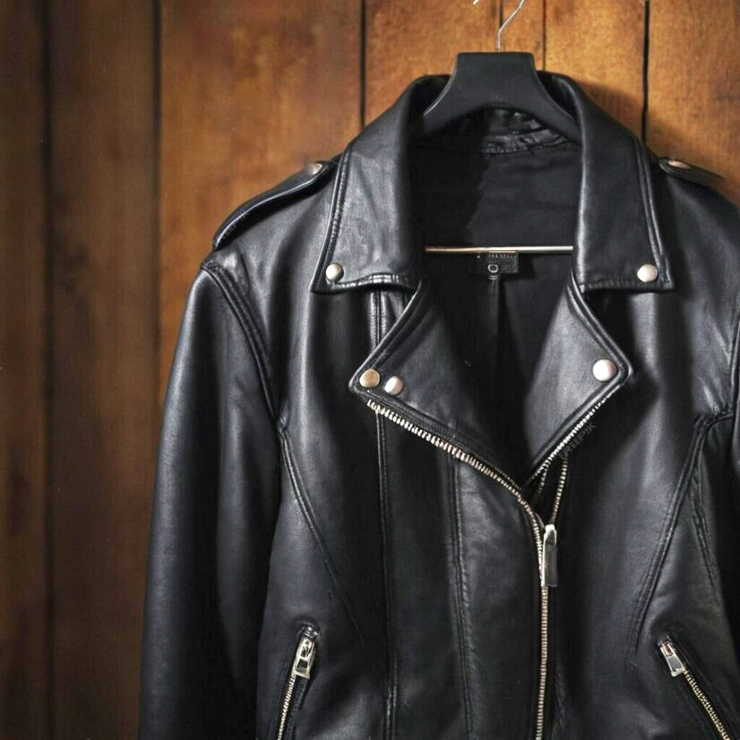 A close-up of a black leather biker jacket hanging on a hanger with silver zippers and snaps.