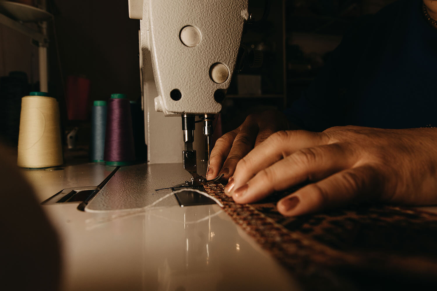 Close-up of hands sewing fabric with a sewing machine, with spools of thread in the background.